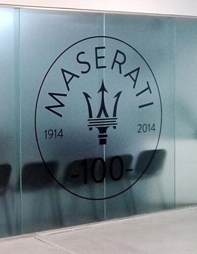 Gigantografía de logotipo de Maserati impreso sobre vinilo esmerilado semi transparente colocado en un panel divisorio de ambiente en un local de venta de automóviles de alta gama.